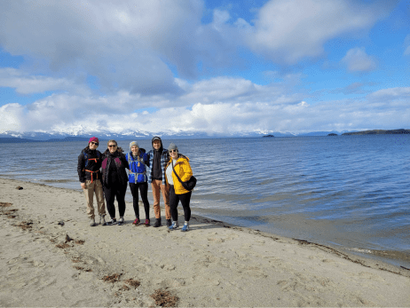 Five students stand along a sandy beach with a large, snow-capped mountain range in the background, along one of Juneau's more scenic hikes.