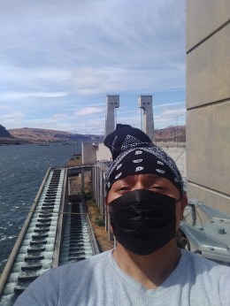 A Native man in a black handkerchief and black face mask is standing above the Big River known as Columbia. He is seen above two rows of salmon fish ladders on a summer day.
