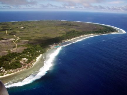 An aerial view of the east coast of the island nation of Nauru. Trees line the perimeter of the island in contrast to the sparsely vegetated interior. The deep blue of the Pacific ocean stretches out in the distance.