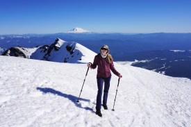 Katie stands atop a snowy mountain peak with a vista in the background.