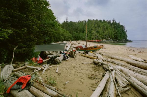 A camp consisting of a small canopy supported by driftwood. Equipment and gear are spread around. The high tide mark is right beside the camp and the dory sits a few hundred feet from the water pulled up onto some large driftwood logs.