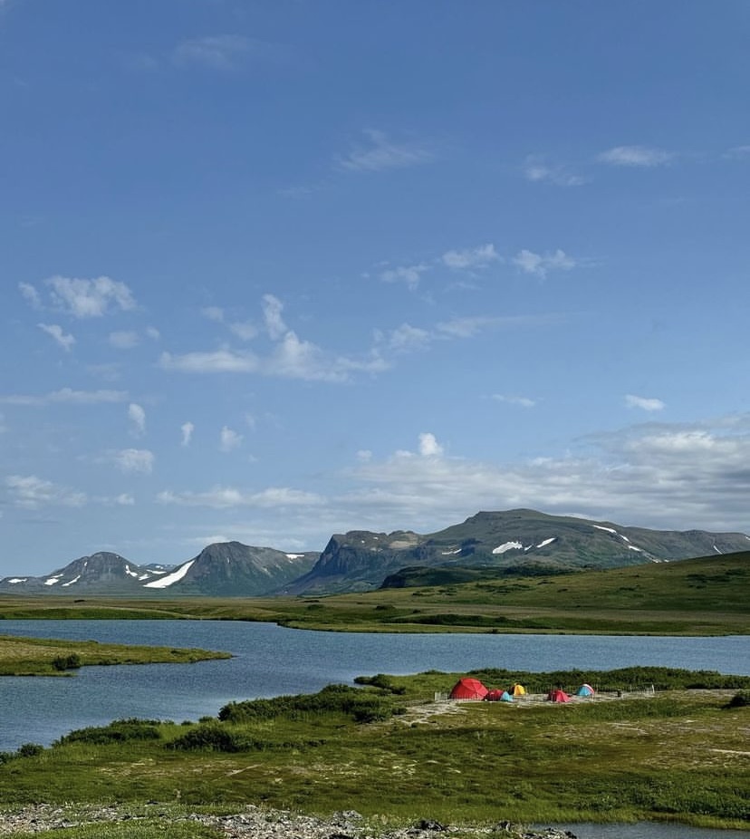 A colorful campground in the distance is situated next to a bright blue lake in Katmai. The reds, yellows, and blues of the tents contrast greatly with the verdant mountains in the background.