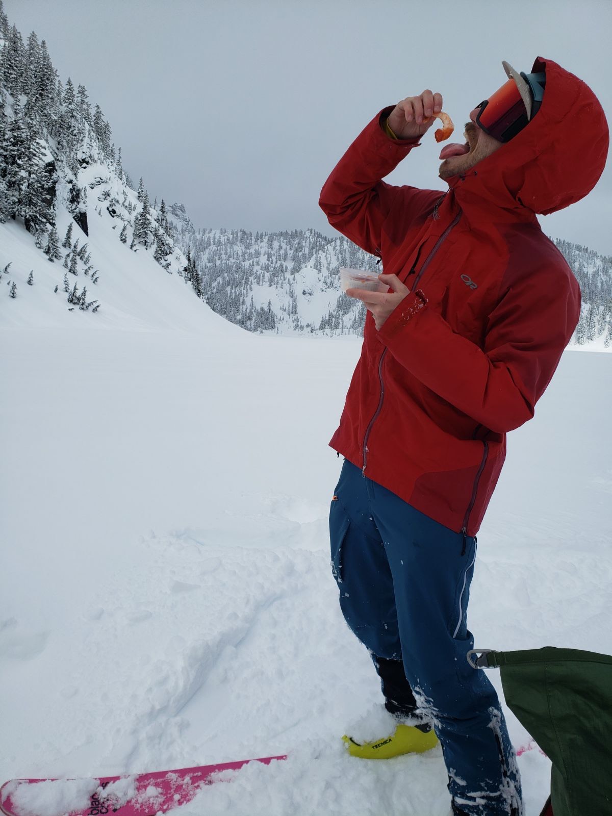 A man dressed in ski touring attire and surrounded by mountains and snow in one hand holds a cooked shrimp with shrimp cocktail sauce over his open mouth and with the other hand holds a shrimp cocktail to-go dish.