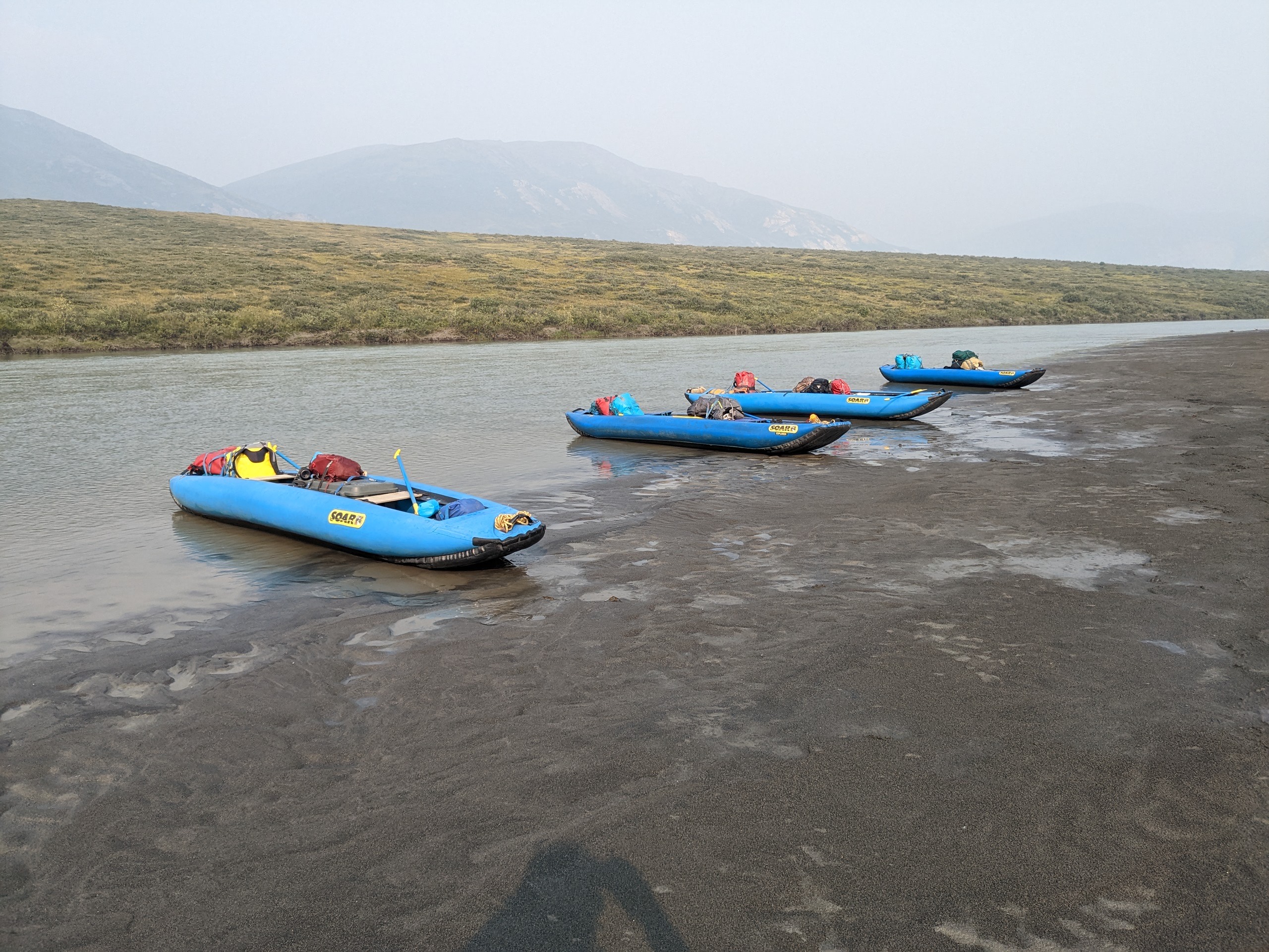 Four bright blue canoes sit on the edge of the riverbank and are fully packed with colorful dry bags, bear drums, clothing, and food.