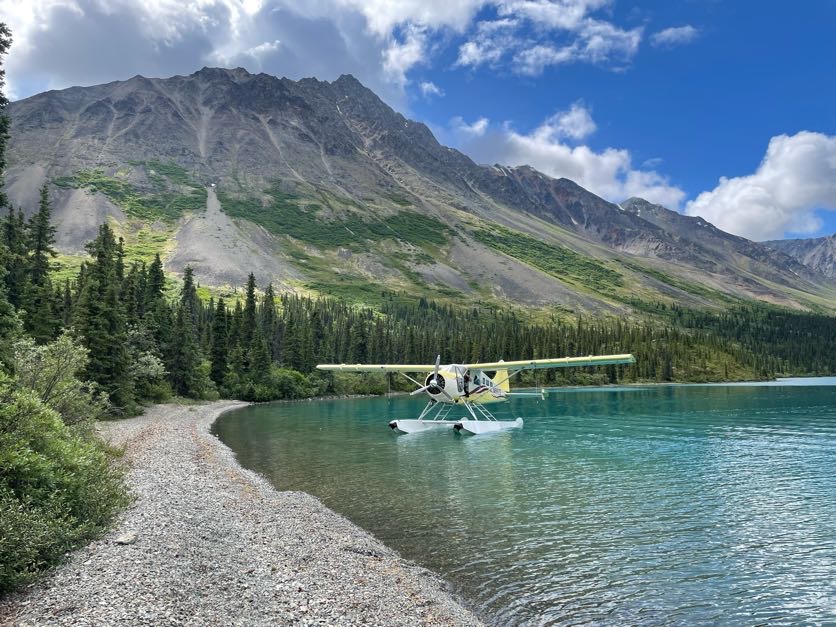 A photo of a yellow float plane resting besides the rocky shore of a crystal blue alpine lake. A tall rocky mountain stretches up behind it.