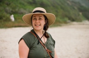 Katalin smiling on a sandy beach in Oregon with greenery on the hill behind her.