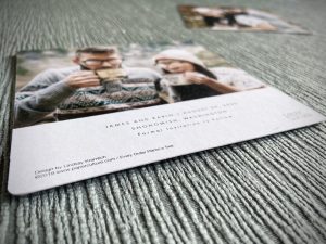 A close up image of a Save the Date for the author's wedding. The card is laying on top of fabric that has a raised vertical stripe weave. The text on the card is indistinguishable due to the angle, but a blurred image of a man and woman wearing glasses and about to drink from mugs is in view.