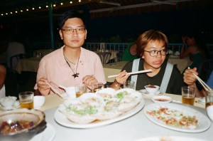 Two people seated at a table with chopsticks in their hands. On the table in front of them are various bowls of food, some containing cooked white rice.