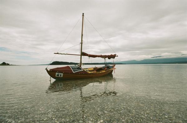 The dory lists to the side in the calm shallows of a shell based beach looking heavy. A solar panel, floatation bags, and a race number on its bow make the boat look official.