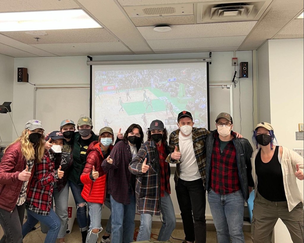 several members of EEL (Engaged Ethnography Lab) line up in front of a projector playing a basketball game; everyone is dressed similarly in flannels and hats to celebrate our advisor's birthday.