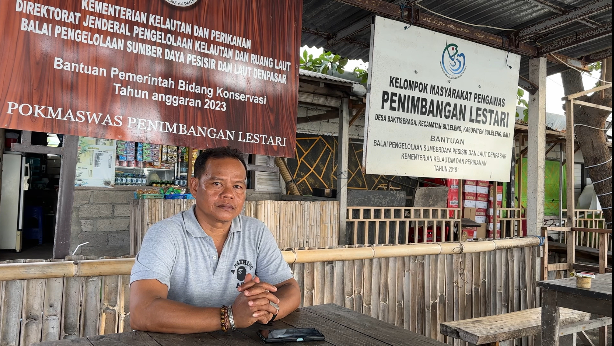 An image of a man in a grey shirt sitting at a wooden table at waterfront food stall.