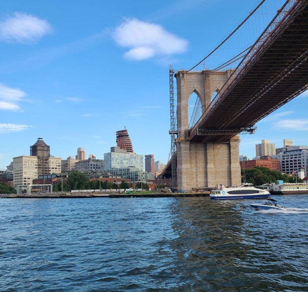A mostly-empty East River with two vessels traveling along the water. Buildings in Brooklyn rise in the background, with the Brooklyn Bridge above.