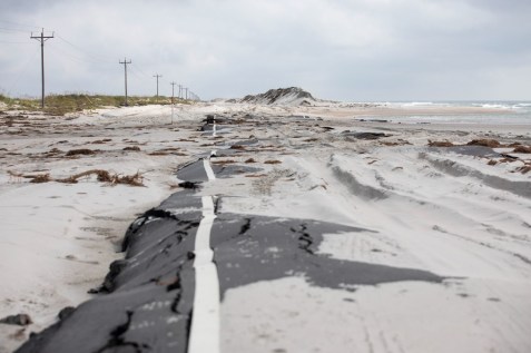 Powerlines, a road, sand dunes and beach extend into the distance. The line of tall dunes is broken and shows the ocean, the beach, and where a storm surge broke through the dunes. The road is buckled and covered in sand. There are tire tracks from cars or trucks in the sand that covers the road.