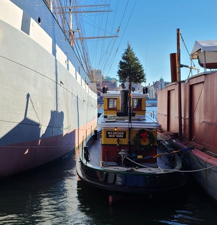 A tug boat with a cheerful red and yellow cabin docked between a much larger ship and a barge. The tugboat is decorated with a Christmas tree and wreath for the holidays.
