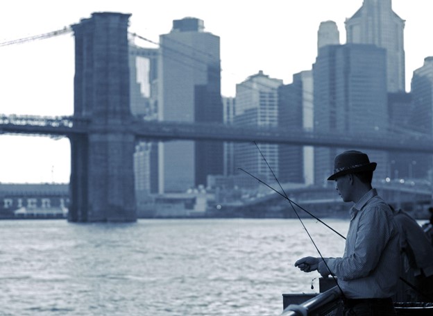 A fisher stands on a pier, holding a fishing line while standing between two fishing poles. The Brooklyn Bridge and Lower Manhattan rise in the background.