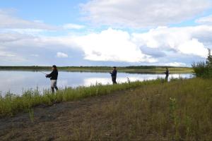 three students stand in some reeds on the bank of a river with fishing rods. The sky is blue with white fluffy clouds and the river is calm.