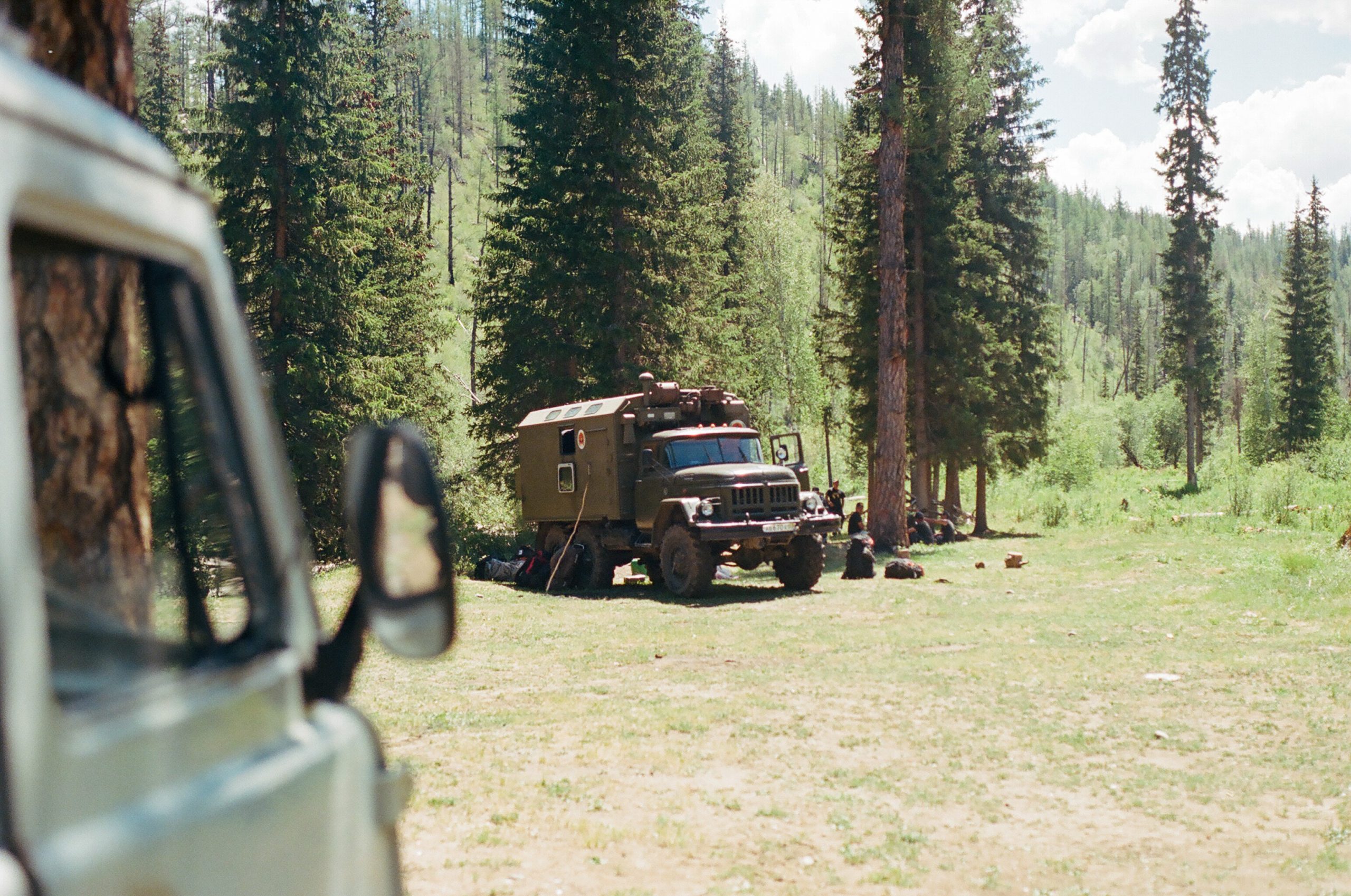 A photo of a giant-knobby-tired 6x6 forest green personnel carrier is taken from behind a small van. Behind the big truck individuals sit under a tree in an apparent meeting.