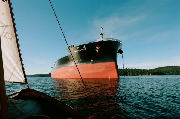 The dory passes close to the looming bow of an oil tanker at anchor. A bright blue sky is contrasted with the deep red hull of the unloaded oil tanker.