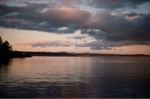 A view across the water from the shore at Friday Harbor Laboratories. The setting sun has lit the island across the channel, and clouds of dark grey and light pink are scattered across a pale sky.