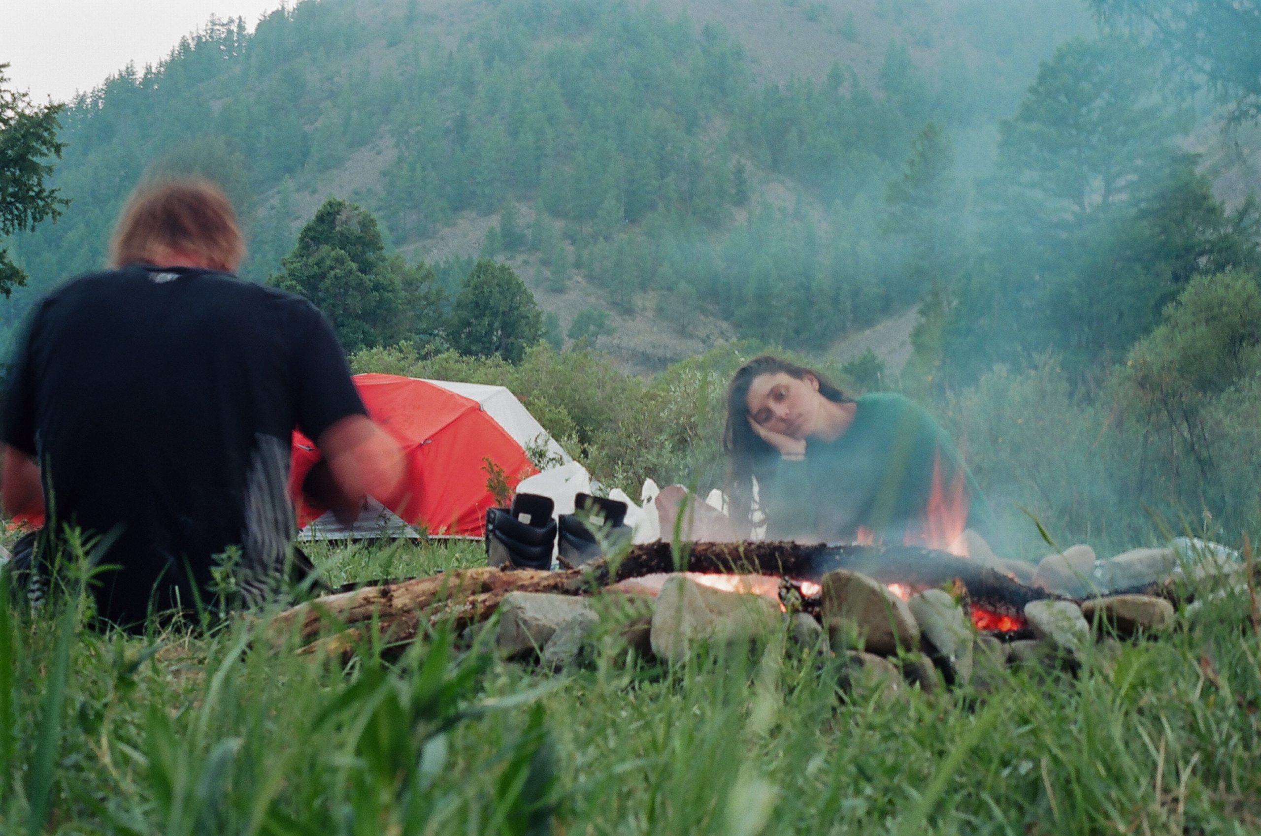 Two people around a campfire with a bright tent in the background. One looks very tired and removed as they look into the fire.