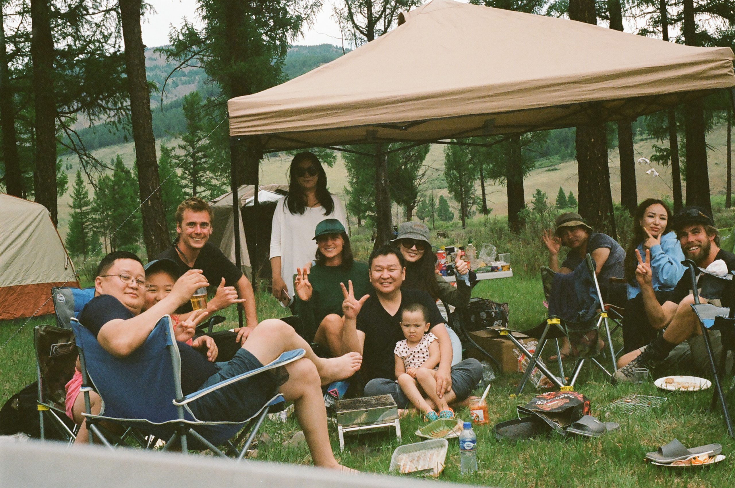 People pose for a photo holding peace signs in crowded campsite in the woods, Food scraps and beer are strewn about and tents populate the background.
