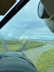 Through the front window of a small airplane, a river stretches below into the distance amongst forest. Just ahead, a rainbow arcs over a small runway.