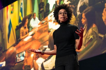 A black woman dressed in black, wearing glasses, and a microphone over her ear is speaking in front of a large theater screen. The picture displayed on the screen is a group of people meeting around a conference room table.