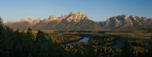 Under a blue sky at sunset, majestic mountain peaks tower above a vibrant pine forest, a sprawling grassy field, and the curving Snake River.