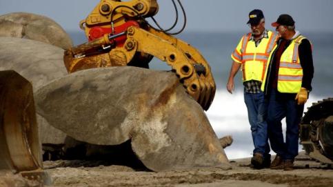 Two people in safety vests on a beach. A large mechanical claw picks up a large rock in the foreground.