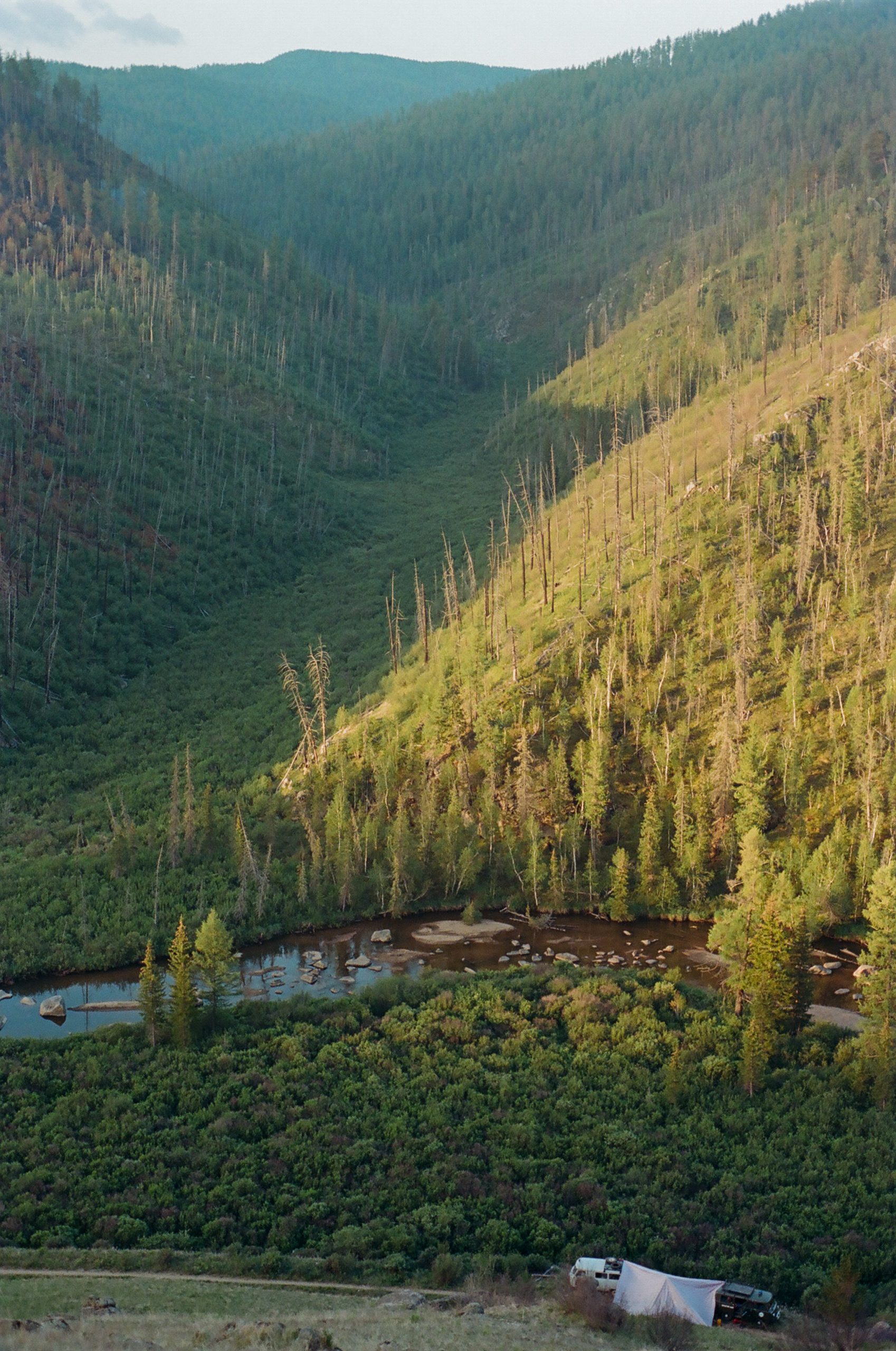 A photo taken from a ridgeline looks up a thickly vegetated green valley. The golden light of a sunrise lights one side of the valley that opens to a still brown river. Looking out of place across the river is a dirt road with two russian 4x4 vans camped along it.
