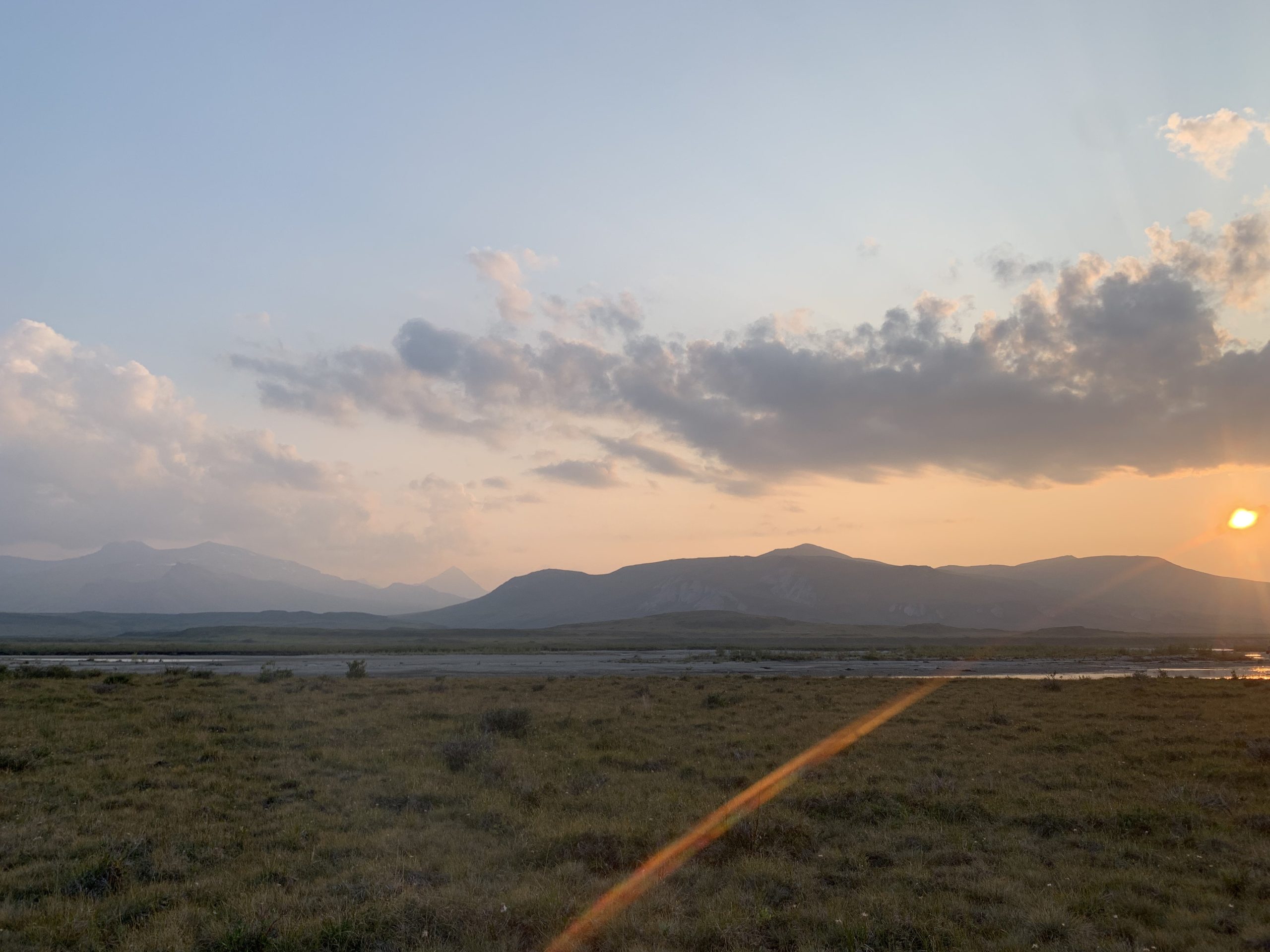 A golden sun sets behind a distant mountain and a ray of sunshine stretches across the Noatak River and a meadow in the foreground, to where the viewer stands. Scattered clouds are clearing and opening up a blue sky.
