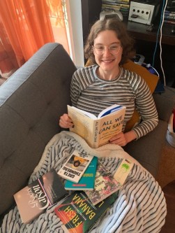 A woman is sitting on a gray couch with an open book in her hands, while many other books lay strewn in her lap.