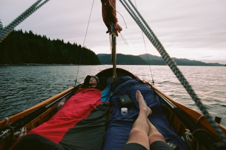 A perspective view with legs outstretched. The author and his very tired friend lay atop sleeping bags while at anchor. Tranquil water around the little boat is slightly blurred giving the impression the world outside the boat is moving while time aboard stands still.