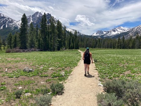 A woman walks on a dirt path through a meadow toward an evergreen forest. The snow-covered Sawtooth Mountains of Idaho stand tall in the background.