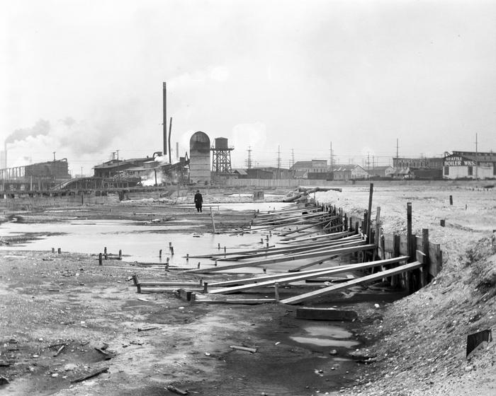 Wood planks hold up soil on the right side of the foreground, while in the center and left the ground remains partially flooded tidelands. In the background a person looks over the construction while behind him are warehouses, a silo, and a water tower.