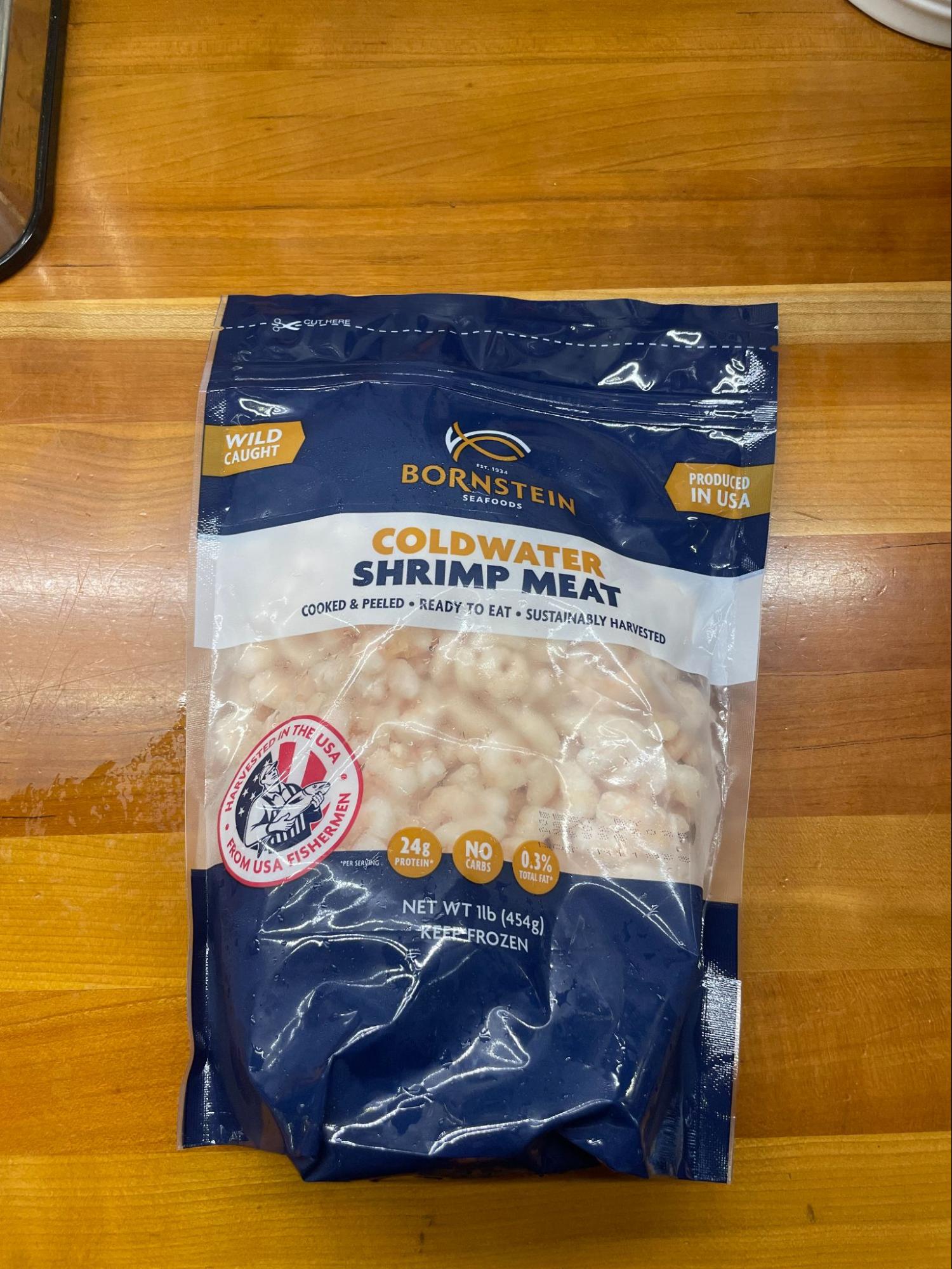 A blue bag of frozen wild caught West Coast pink shrimp on a wood countertop. The bag reads " Bornstein seafood: Coldwater Shrimp Meat - cooked & peeled - ready to eat - sustainably harvested" and has a logo with a fisherman holding a fish with the words: "Harvested in the USA, from USA fishermen."