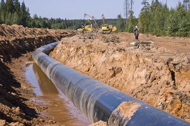 image shows the construction of a pipeline, a large black pipeline surrounded by damaged land and dirty water with construction in the background.