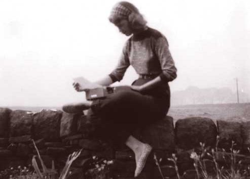 A black and white photo of Sylvia Plath as a young woman perched on a low stone wall with a typewriter on her lap in September 1956.