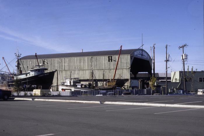 A long grey-green warehouse photographed from its parking lot. A fishing vessel is dry-docked on the left side of the photo for repairs.