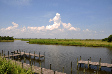 Wooden docks sit along a river, marsh grass, blue sky, and puffy clouds are visible in the background.