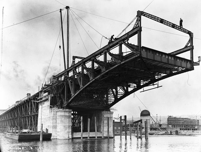 Half of a drawbridge hangs over a river, with workers bending over different parts of the bridge.