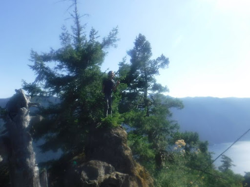 A happy figure flashing a peace sign sands on a ledge high above the mountain valley and lake.