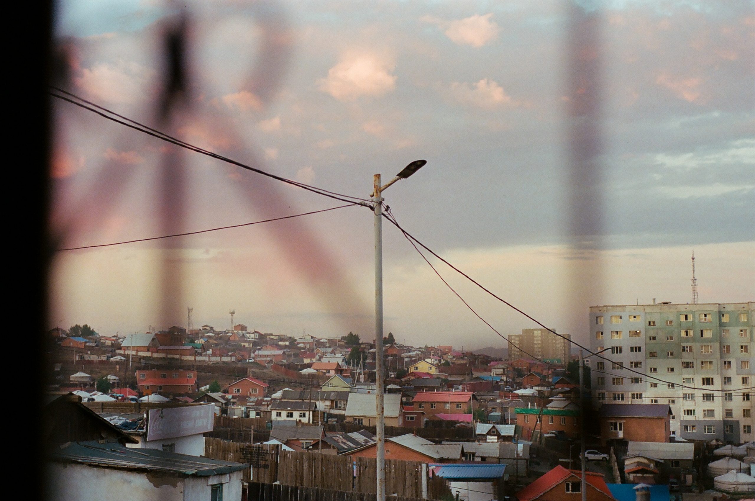 A photo taken from behind a fence looks out at a sunset sky over brightly colored roofs and the occasional white yurt or ger. Two multistory apartment buildings interject amidst the densely populated scene.