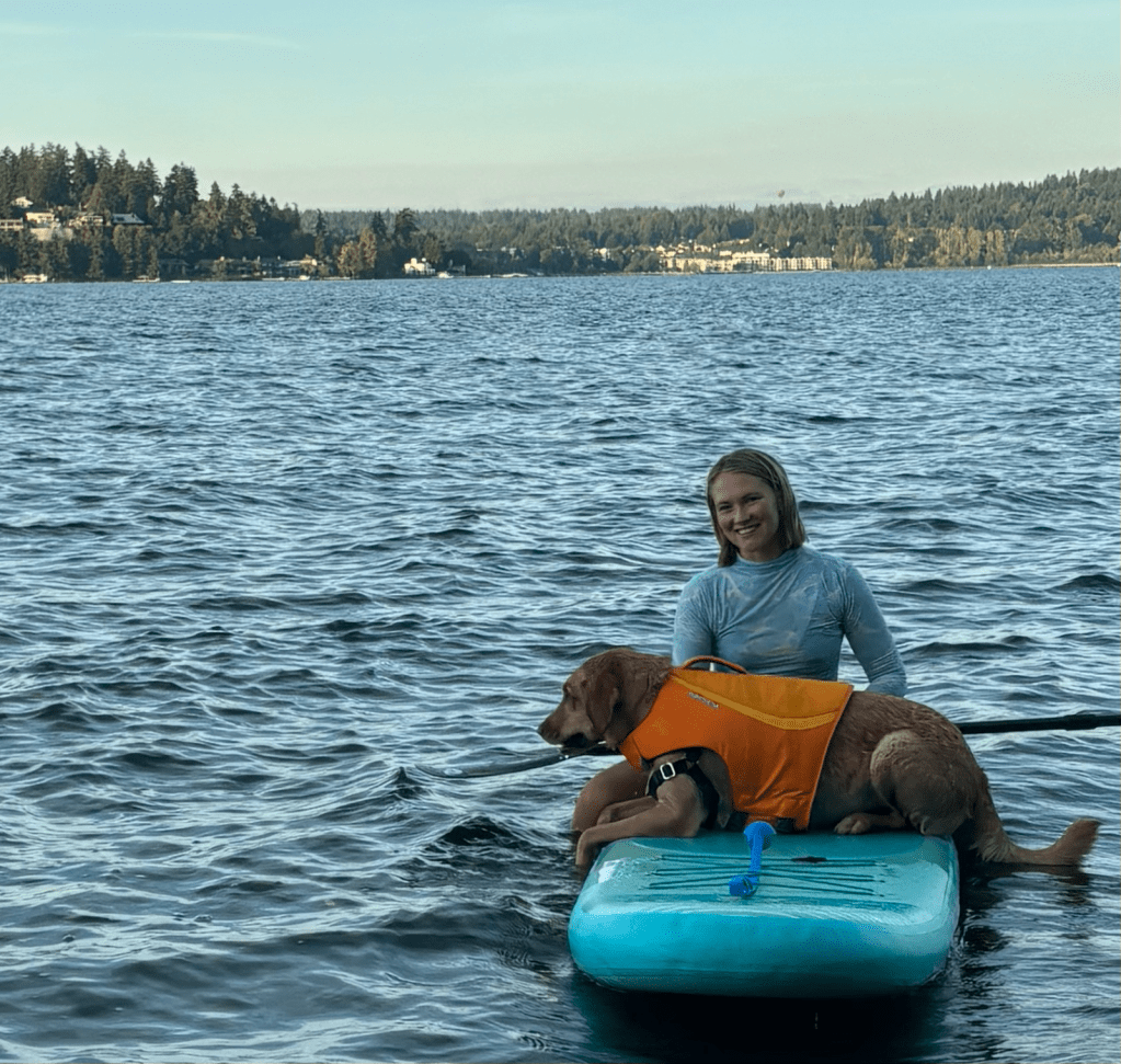 Alt text: A photo of Amie Kusch sitting atop a paddleboard on a lake with a large dog wearing an orange life jacket.
