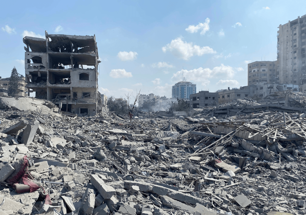 Alt text: A field of rubble is all that's left of a few flattened concrete buildings. In the background, bombed out shells of apartment buildings are visible as far as the eye can see.