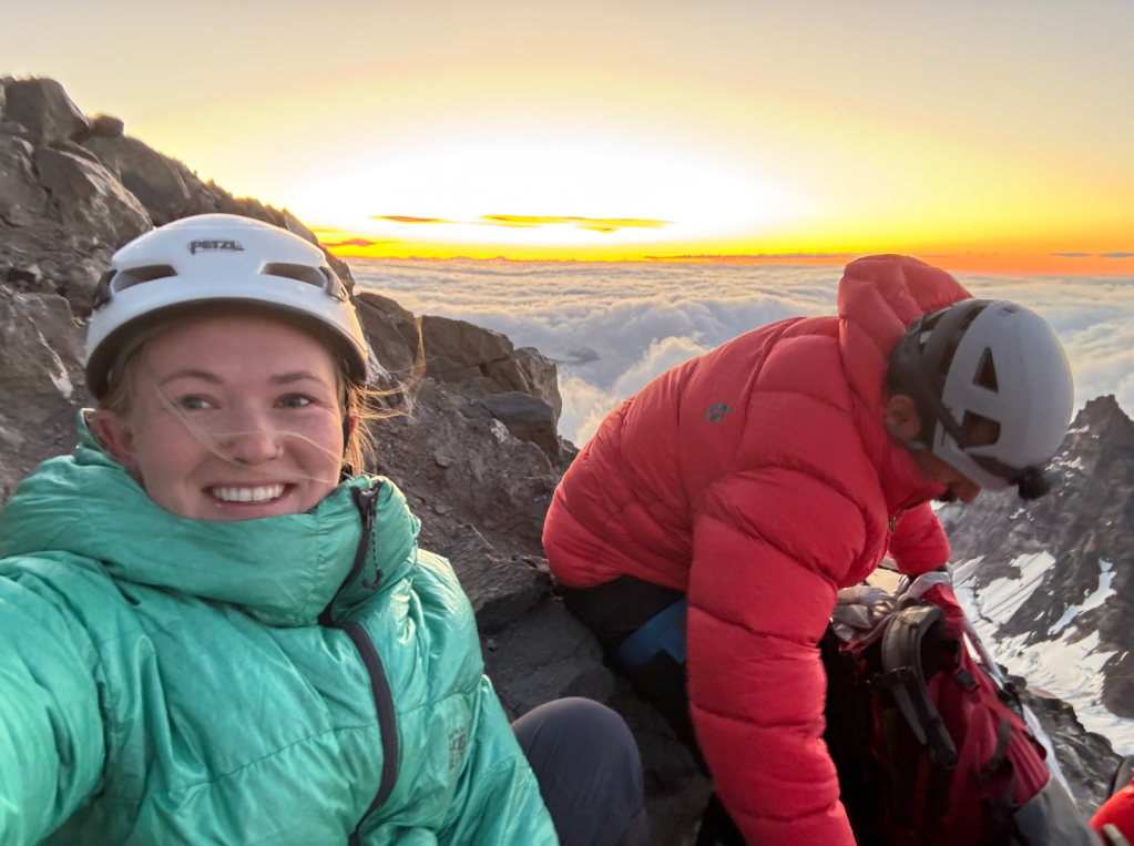 Alt text: The Author, Amie, and another climber sitting on rocks at sunrise above the clouds. Amie is smiling at the camera in a blue jacket and helmet; the person on the right wears a red jacket and adjusts a backpack.