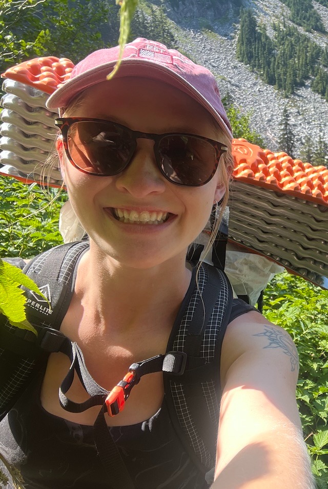Alt text: A selfie of Anna Lewis equipped with backpacking gear in a mountainous setting.