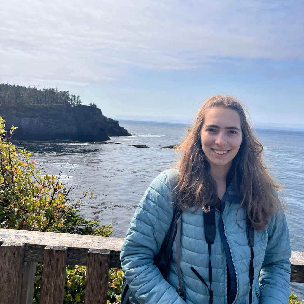 Alt text: a photo of Bea Pickett standing at a coastal viewpoint with ocean and rocky headlands in the background.