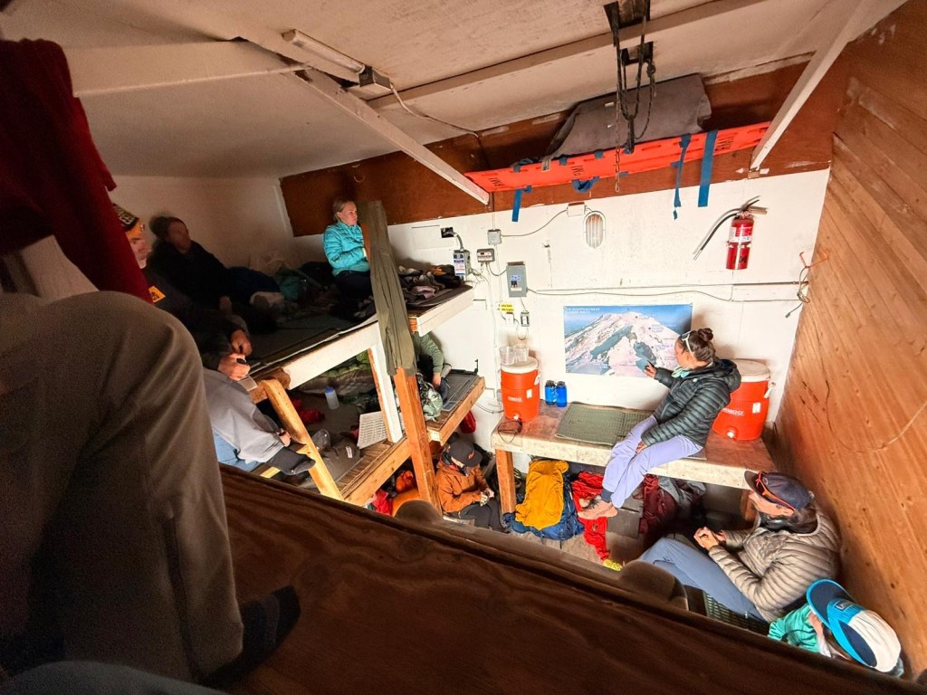 Alt text: Inside a small wooden hut with several of the climbers sitting on bunks and benches. A guide points to a large map on the wall of Mount Rainier while others listen. Gear is scattered around the bunk.