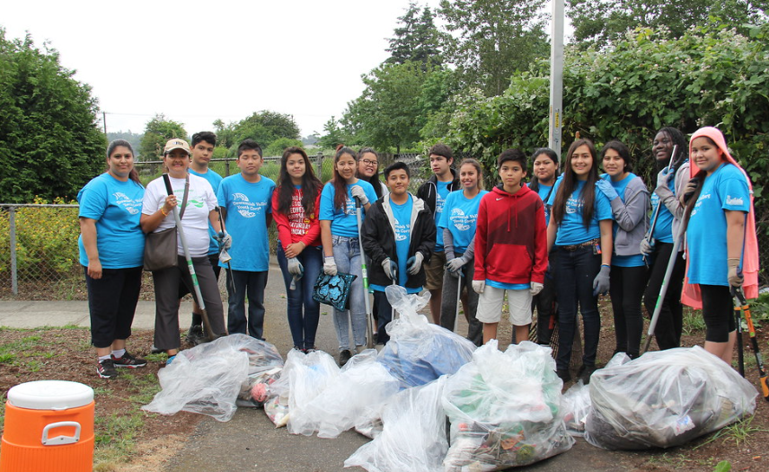 Alt text: The Duwamish Valley Youth Corp participates in a clean-up event. A group of people wearing light blue shirts stand behind bags of trash collected during the clean-up event. 